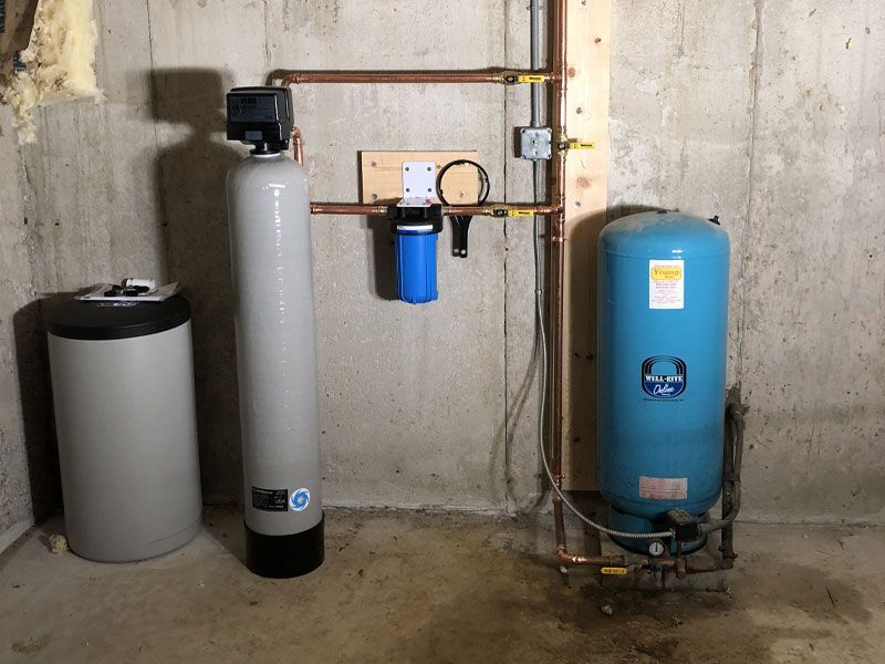 Basement utility room with water softener, filtration tanks, and plumbing pipes against a concrete wall