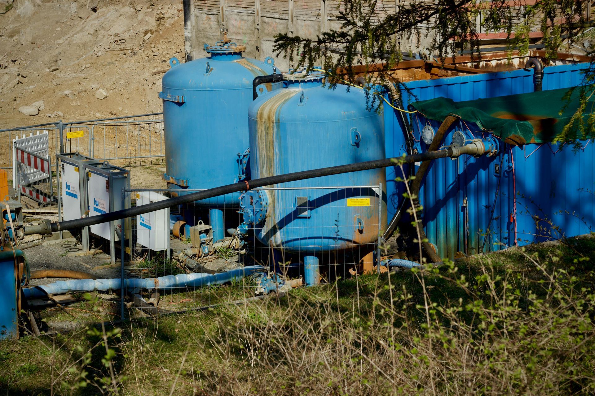 Blue industrial tanks and pipes beside a rocky hillside with grass in the foreground