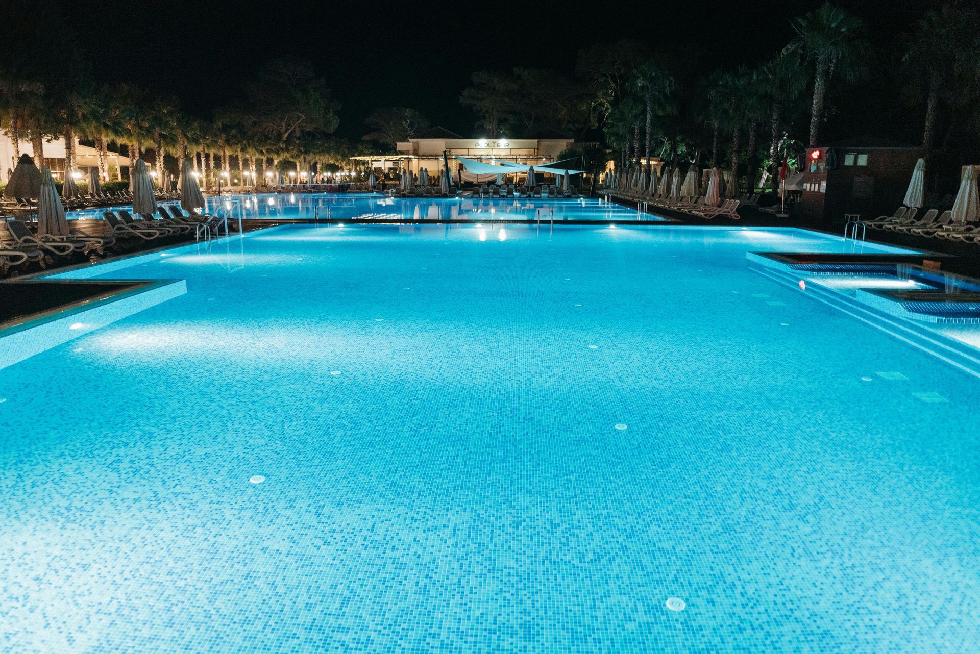 Illuminated outdoor swimming pool at night with bright blue water and lounge chairs along the sides