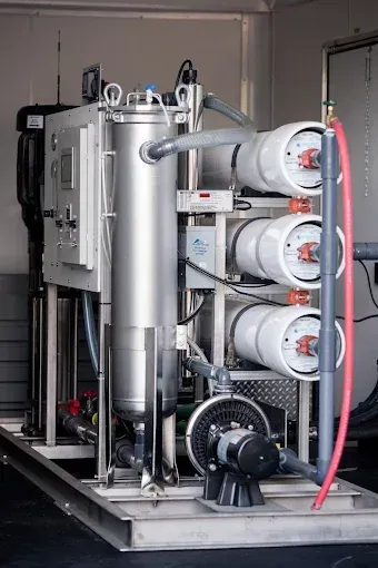 Industrial water filtration unit with stainless steel tanks, white filter housings, and red hose in a utility room