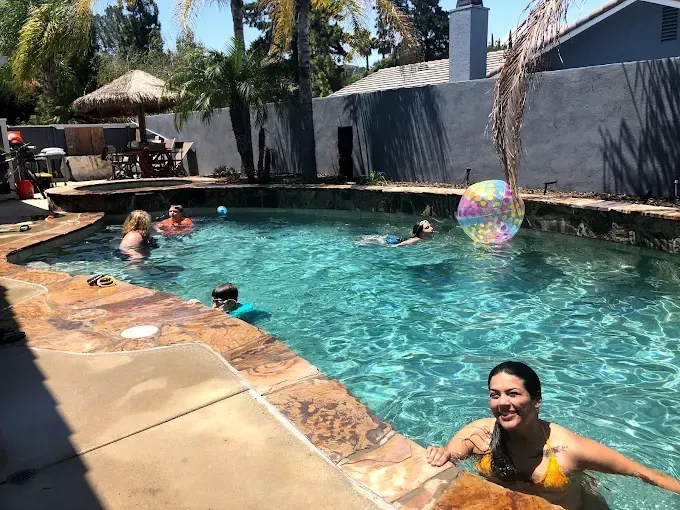People swimming in a backyard pool on a sunny day, with a colorful beach ball and patio seating nearby