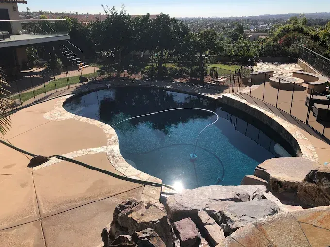 Backyard swimming pool with tan patio, rocks, and trees in sunlight