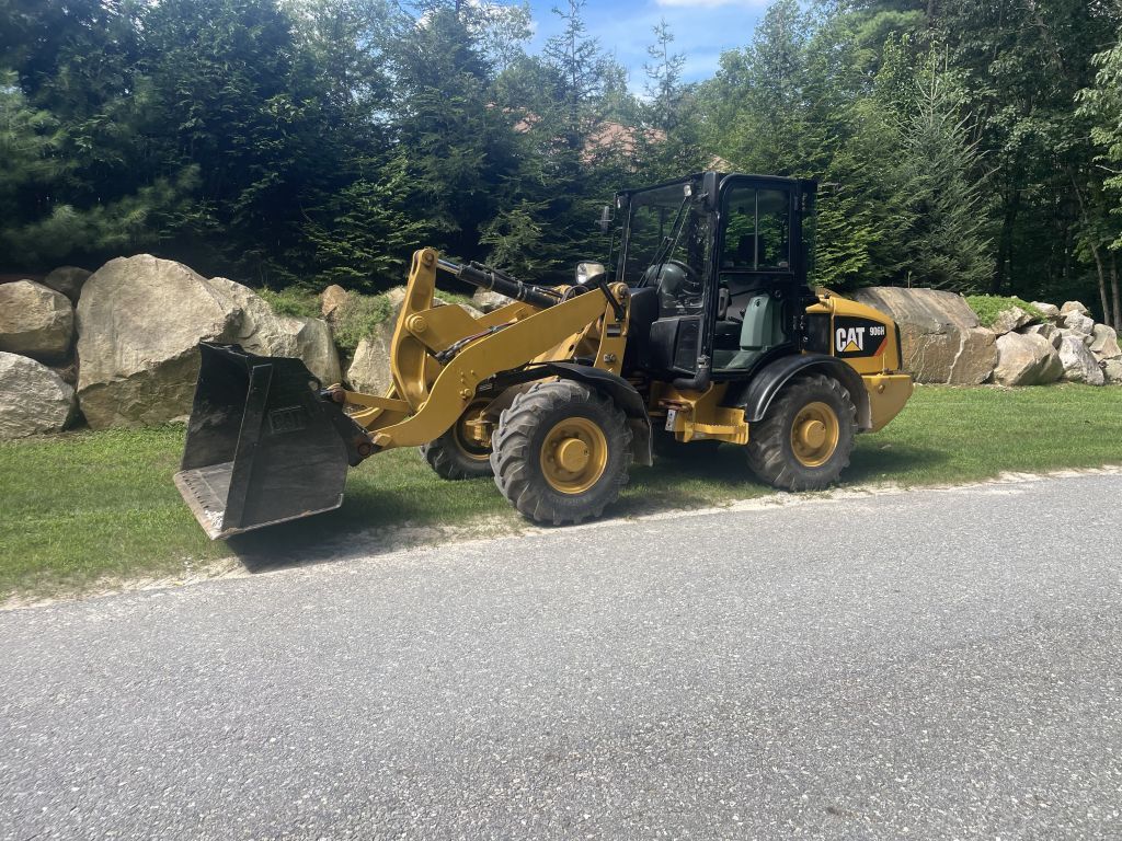 Yellow Caterpillar wheel loader parked on a gravel path near large rocks and trees.