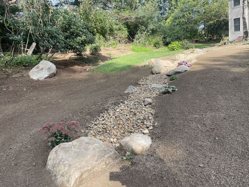 Grey house exterior with steps leading to a dark blue door, surrounded by greenery and grass.