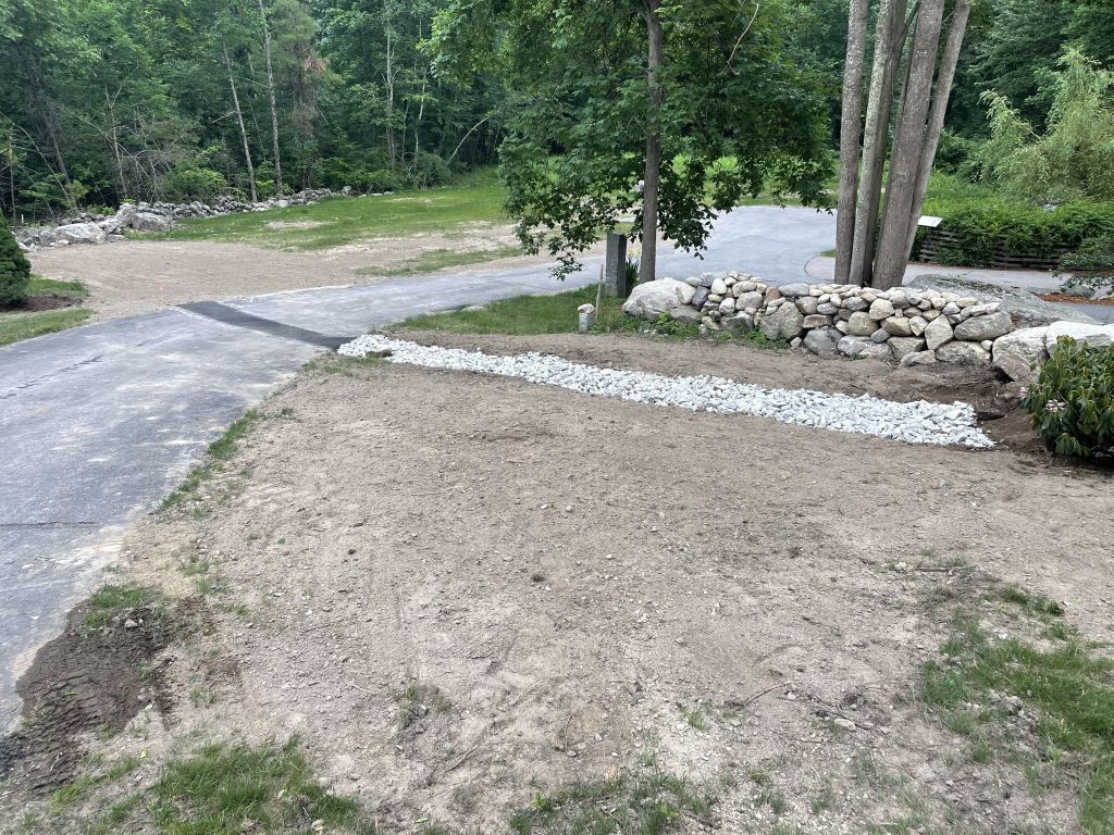 Trench dug alongside a yellow house, construction equipment visible in the background, daytime.