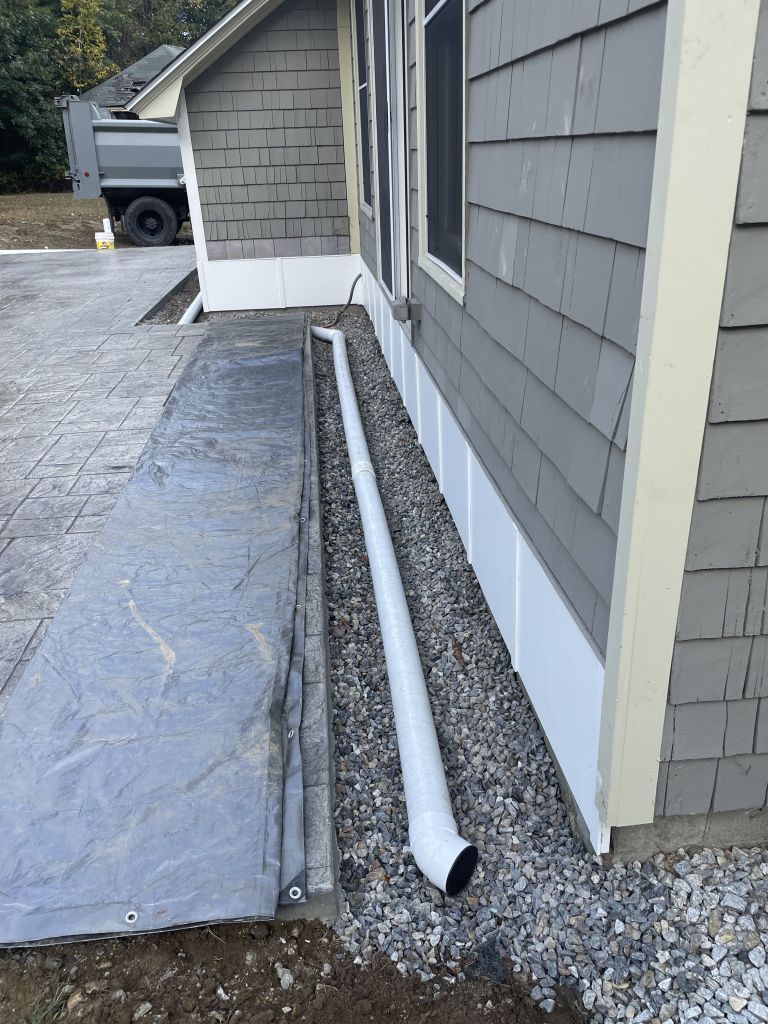 Excavation beside a yellow house, exposing foundation, gravel, and drainage pipe.