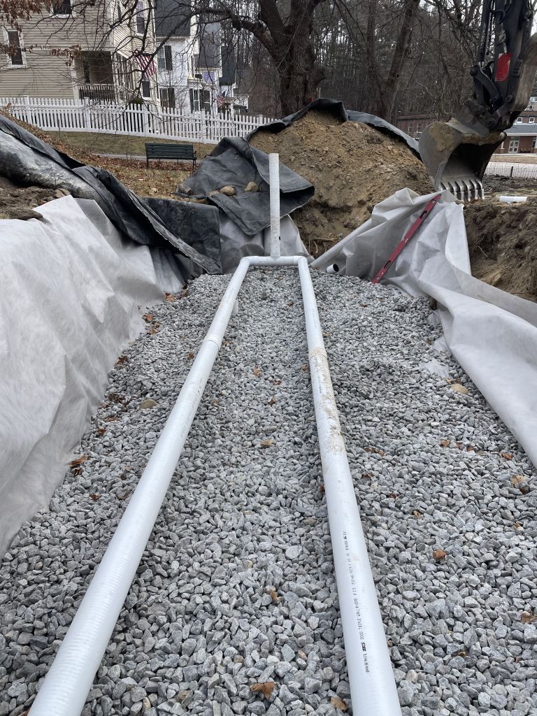 Construction site, septic system installation. Black pipes, white elbows, sand, dirt, and a tree in the background.