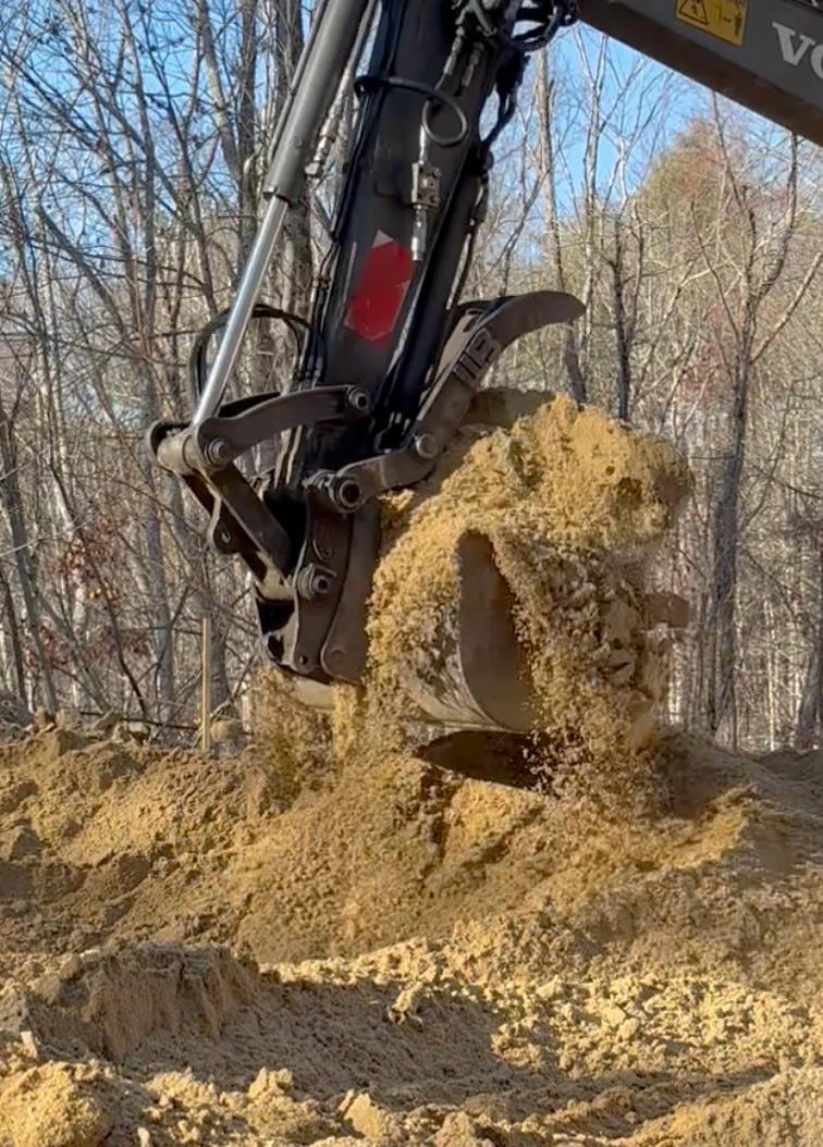 Gravel patio with a border, grass, and a large bush in front of trees.