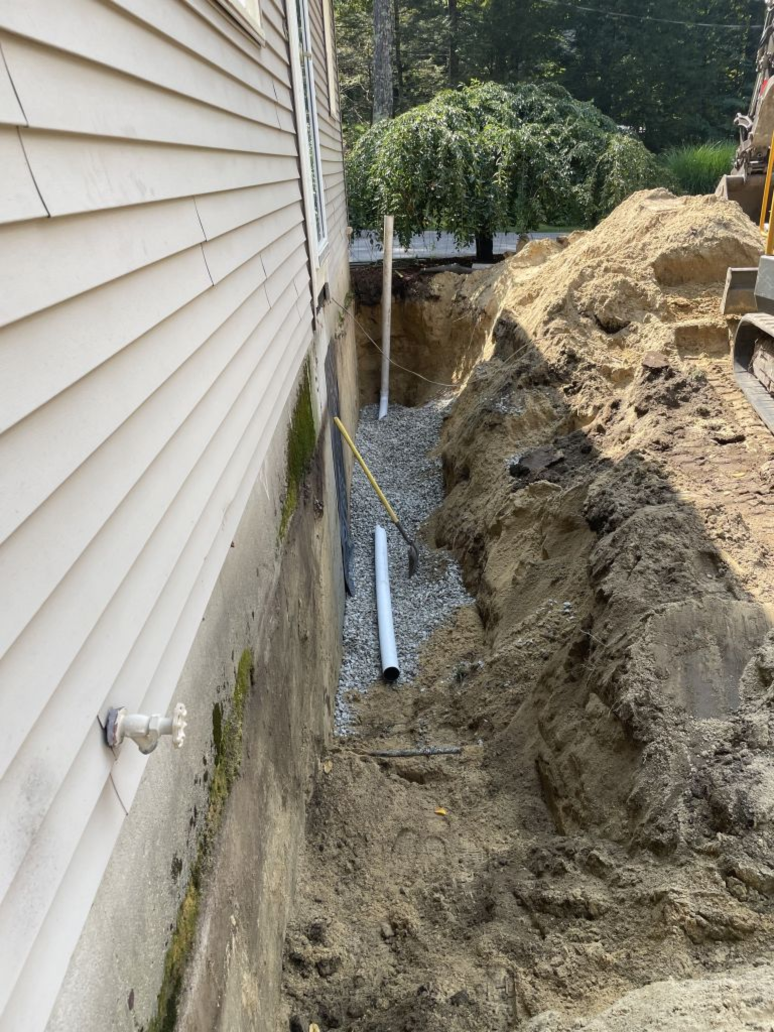 Trench dug beside a house wall. White pipes and gravel are visible within the trench.