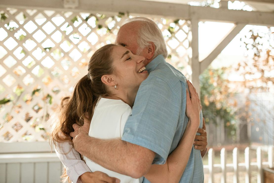 Two people embracing and smiling in a sunlit outdoor gazebo with a white lattice background