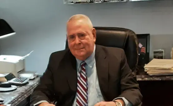 Man in suit seated at desk, smiling. Desk with calculator, paper, and lamp in office setting.