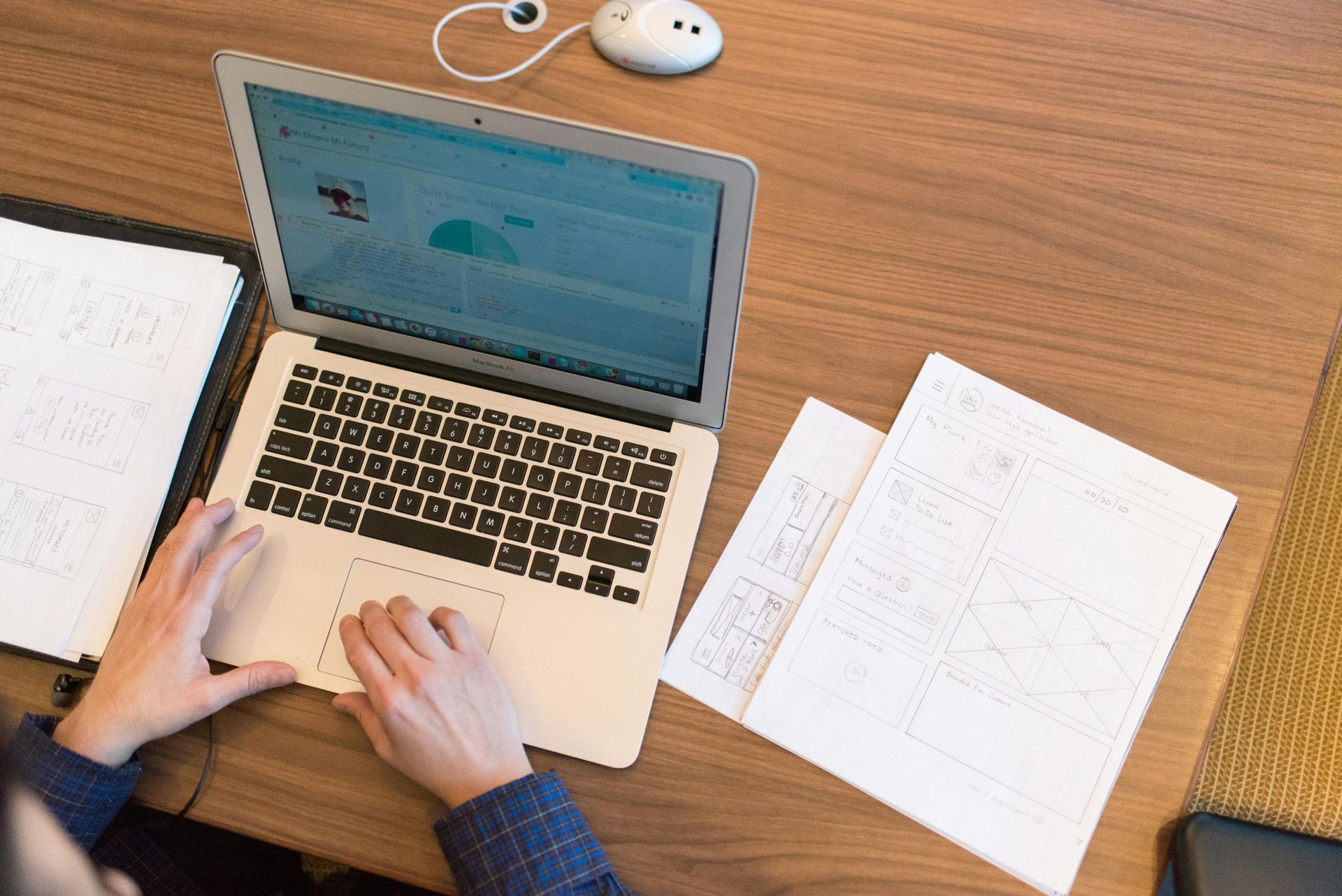 Person using a laptop at a desk with papers. A mouse and notebook are also visible.
