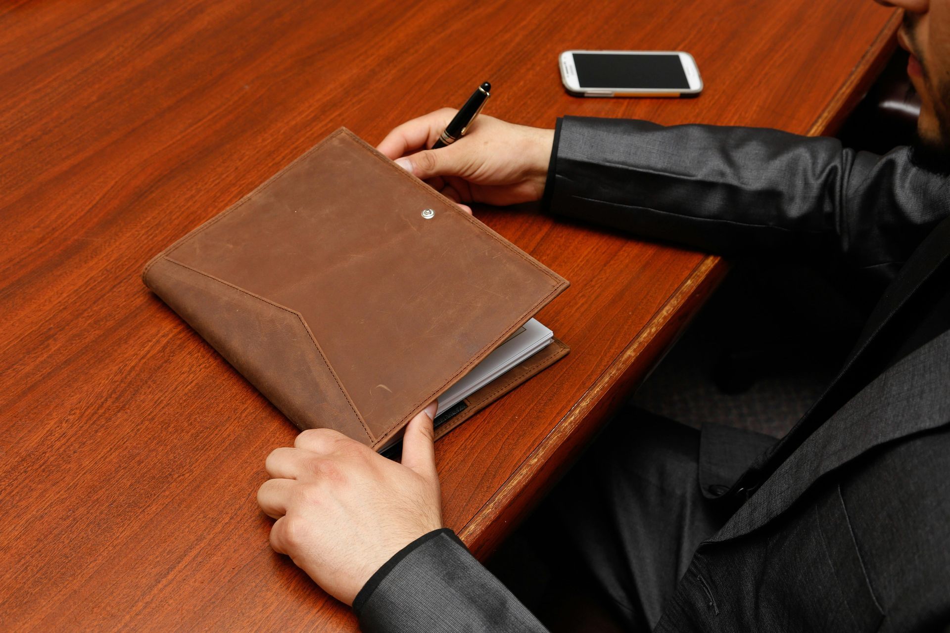 Person in suit opening a brown leather notebook on a wooden desk with a pen and phone.
