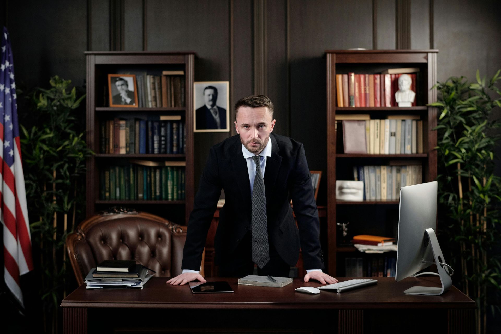 Man in suit standing at desk in a wood-paneled office; American flag, bookshelves, and computer monitor in background.