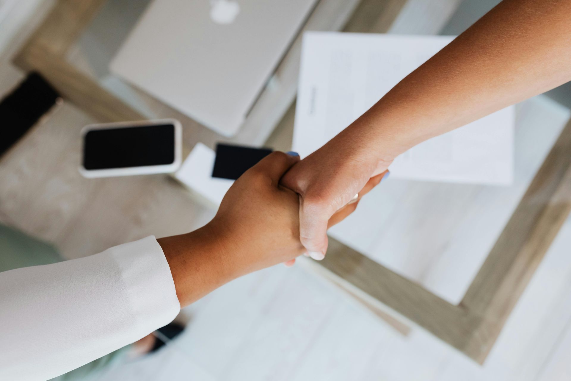 Two people shaking hands over a table with laptop and phone.
