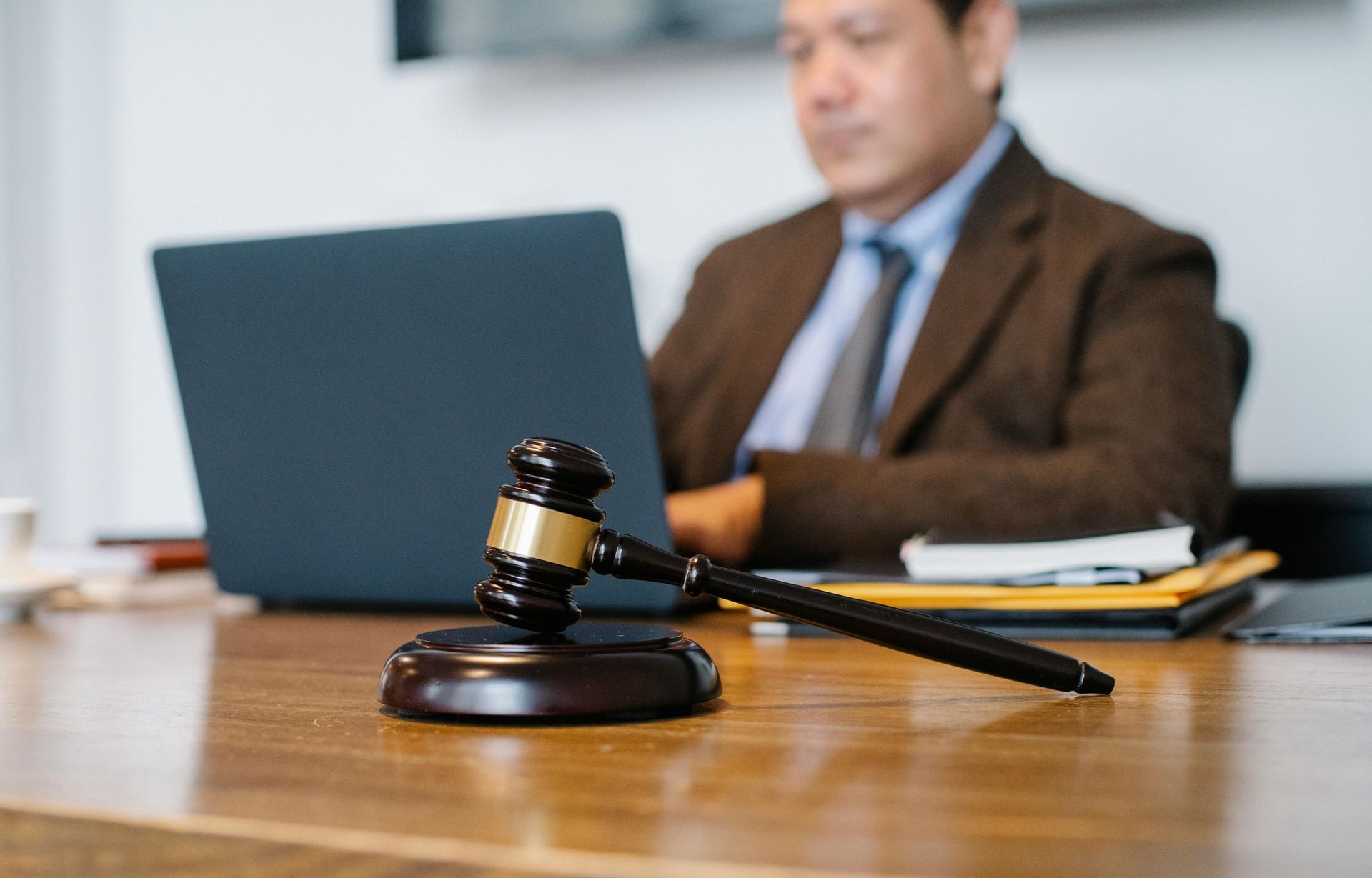 Gavel on desk in foreground, man in suit using laptop in background.