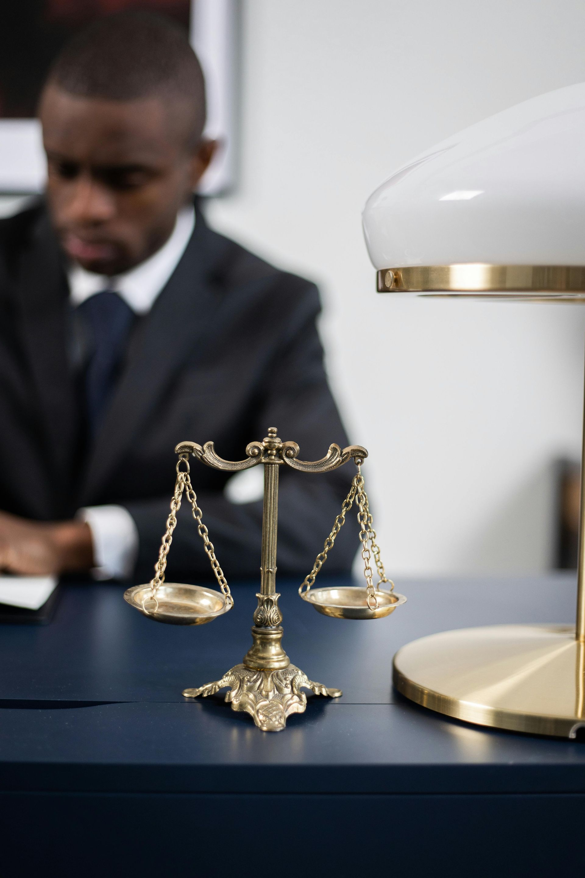 Gold scales of justice on a desk; a man in a suit is blurred in the background. A desk lamp is to the right.