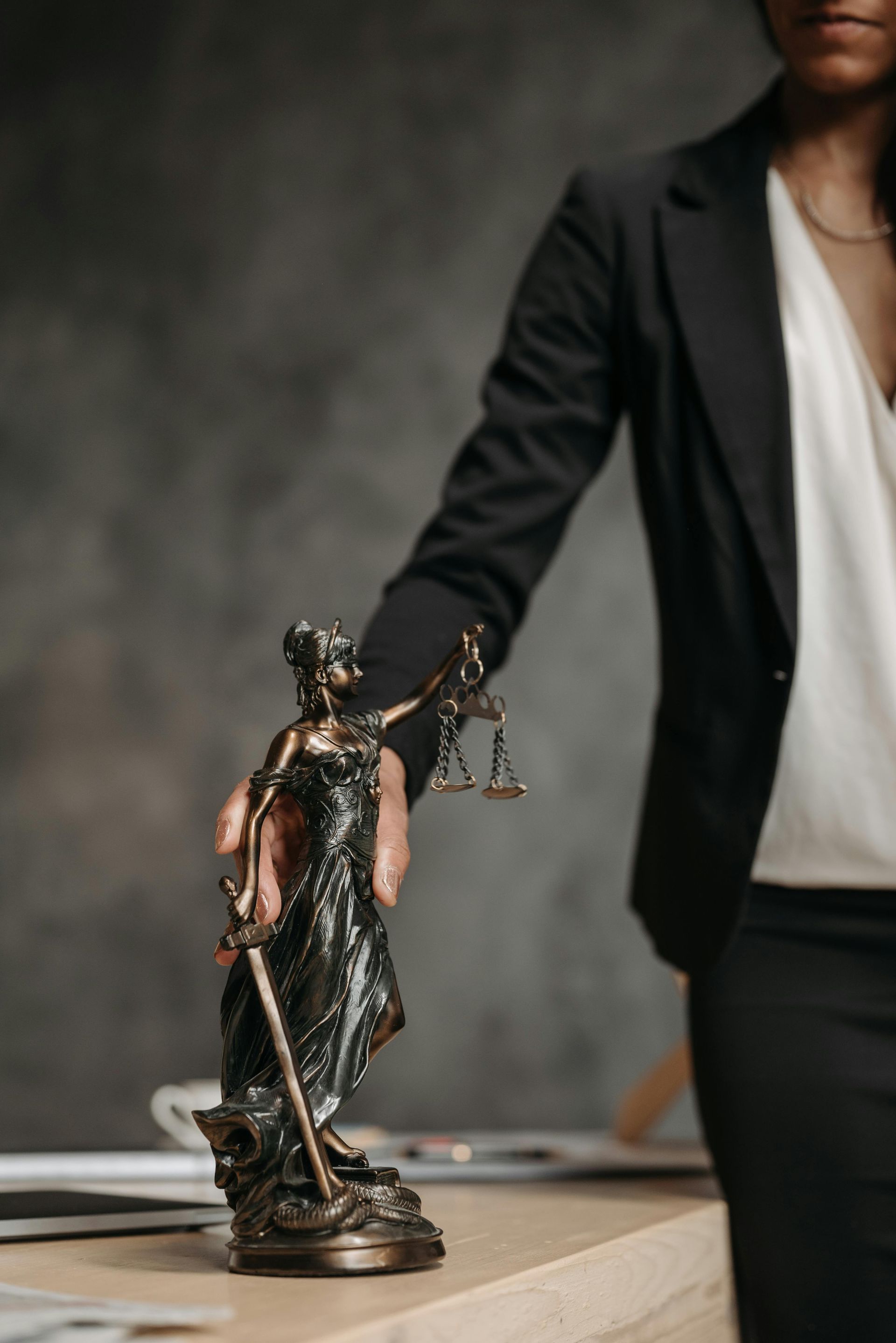 Woman in suit, holding statue of Lady Justice. Scales and sword. Dark background.