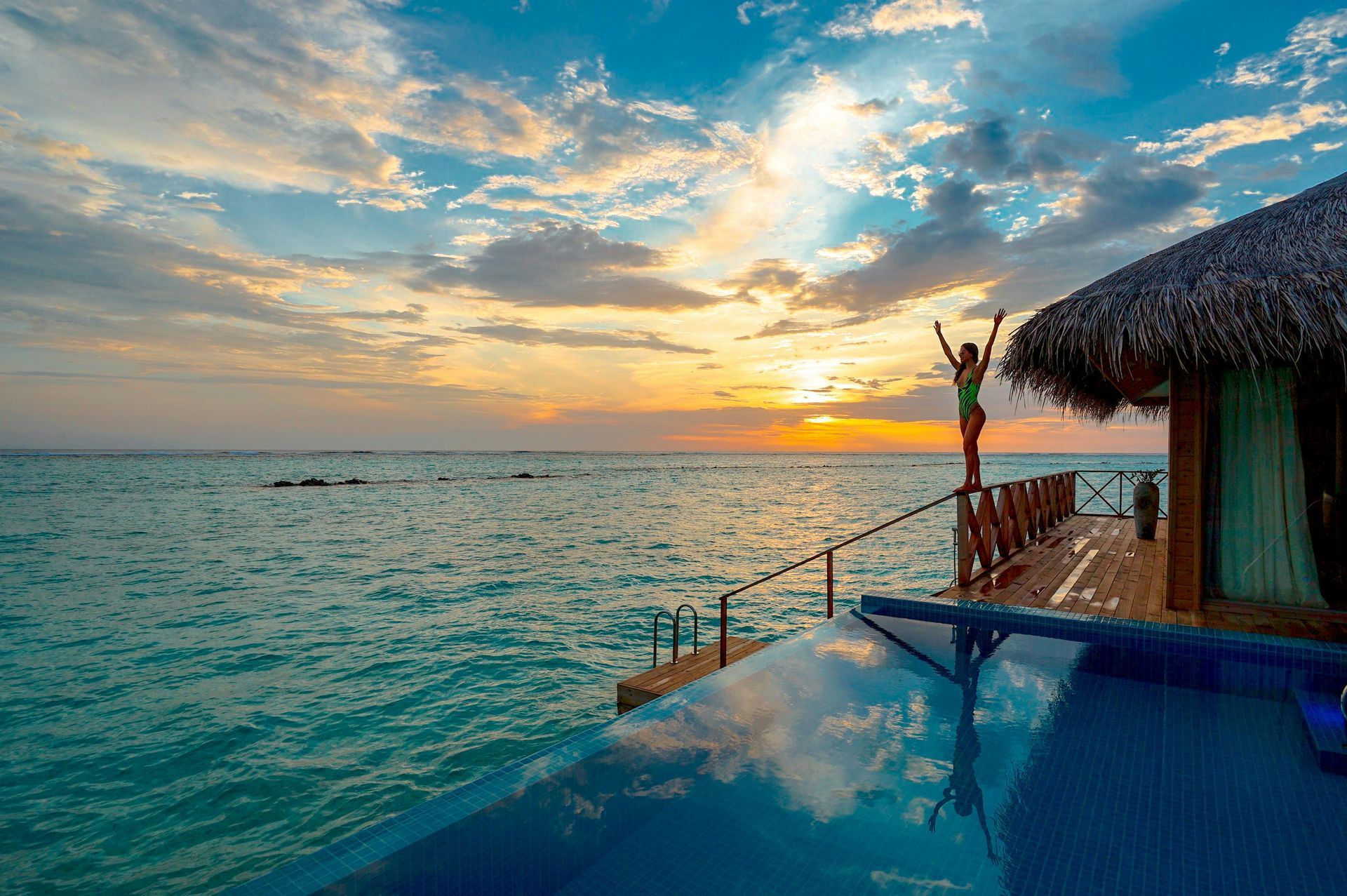 Woman with arms raised at sunset, standing on wooden deck with thatched roof. Pool overlooks ocean.