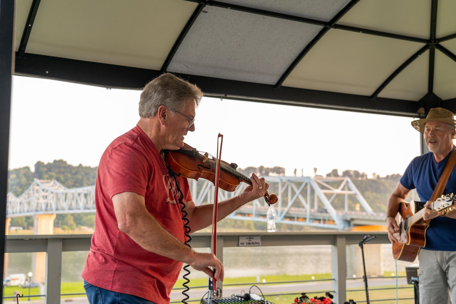 Two men are playing violins and guitars under a canopy.
