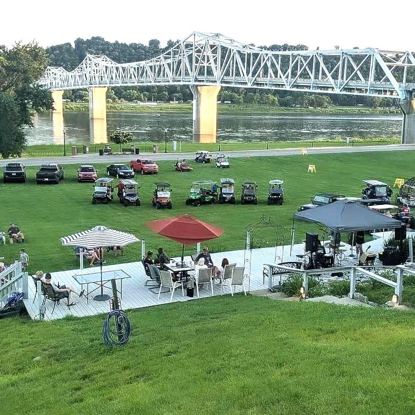 A group of people are sitting at tables under umbrellas in a field with a bridge in the background.