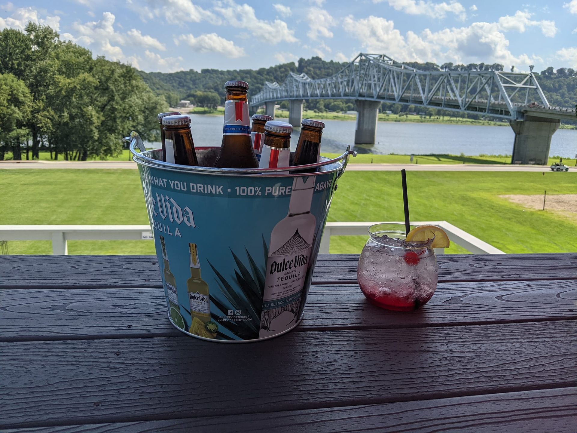 A bucket of beer and a drink on a table with a bridge in the background.