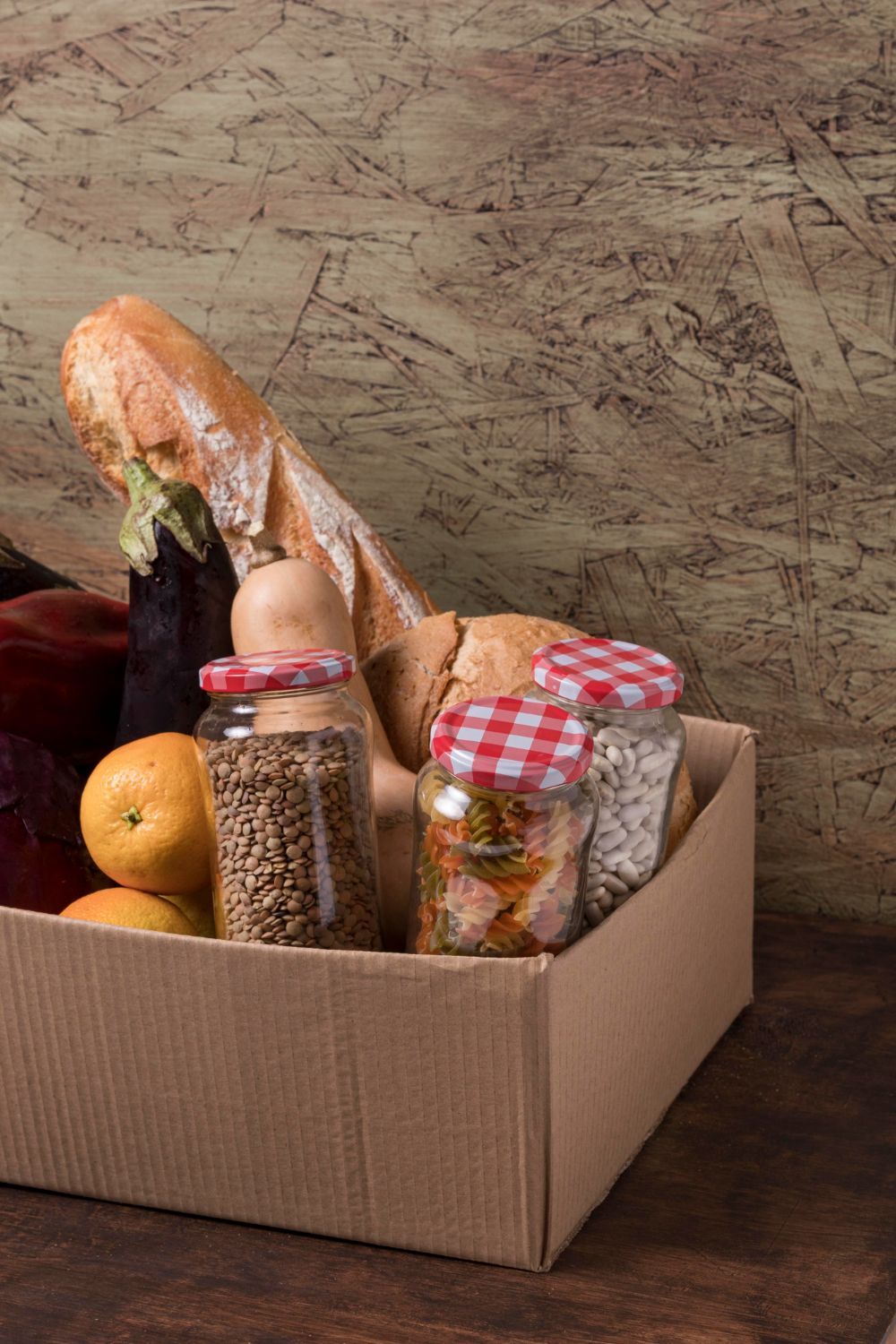 bandejas de almacenamiento Caja de cartón llena de comida: baguette, tarros de cereales y especias, y fruta. Sobre un fondo de madera veteada.