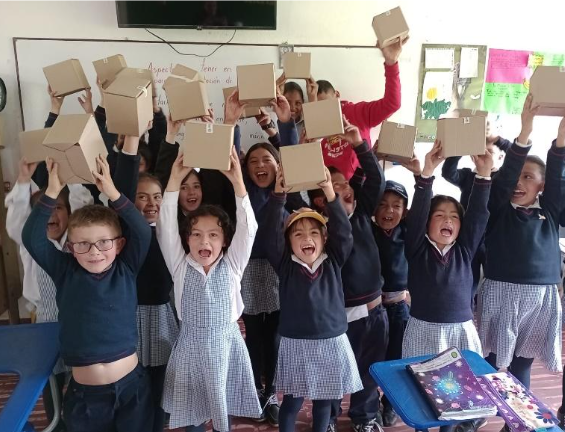 Un grupo de escolares emocionados, uniformados, levanta cajas de cartón en un aula. Todos sonríen.