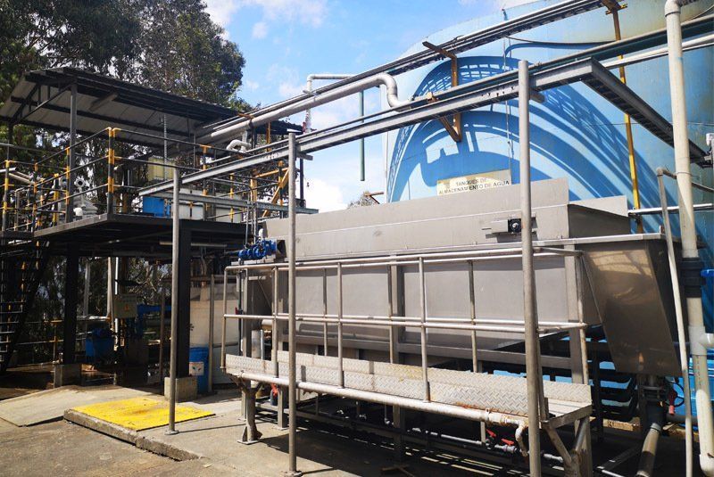 Una instalación de tratamiento de agua con tanques, tuberías y pasarelas metálicas, ubicada al aire libre bajo un cielo parcialmente nublado.