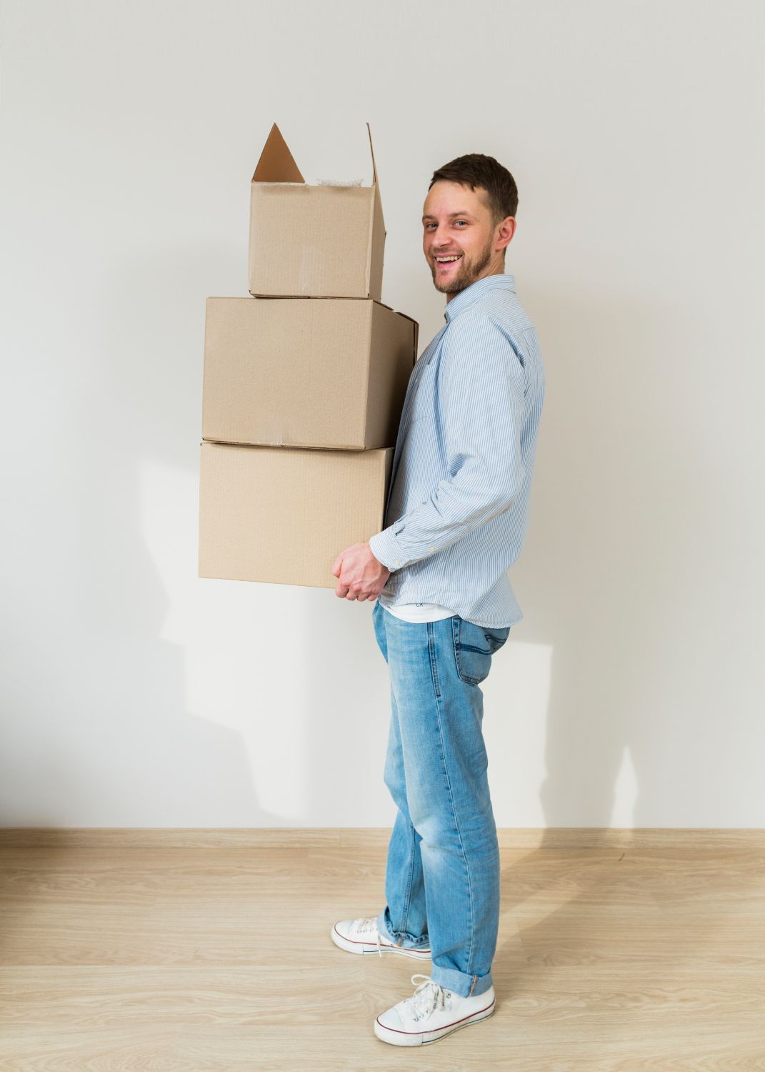 Hombre sosteniendo una pila de tres cajas de cartón, sonriendo, de pie en una habitación con paredes claras y pisos de madera.