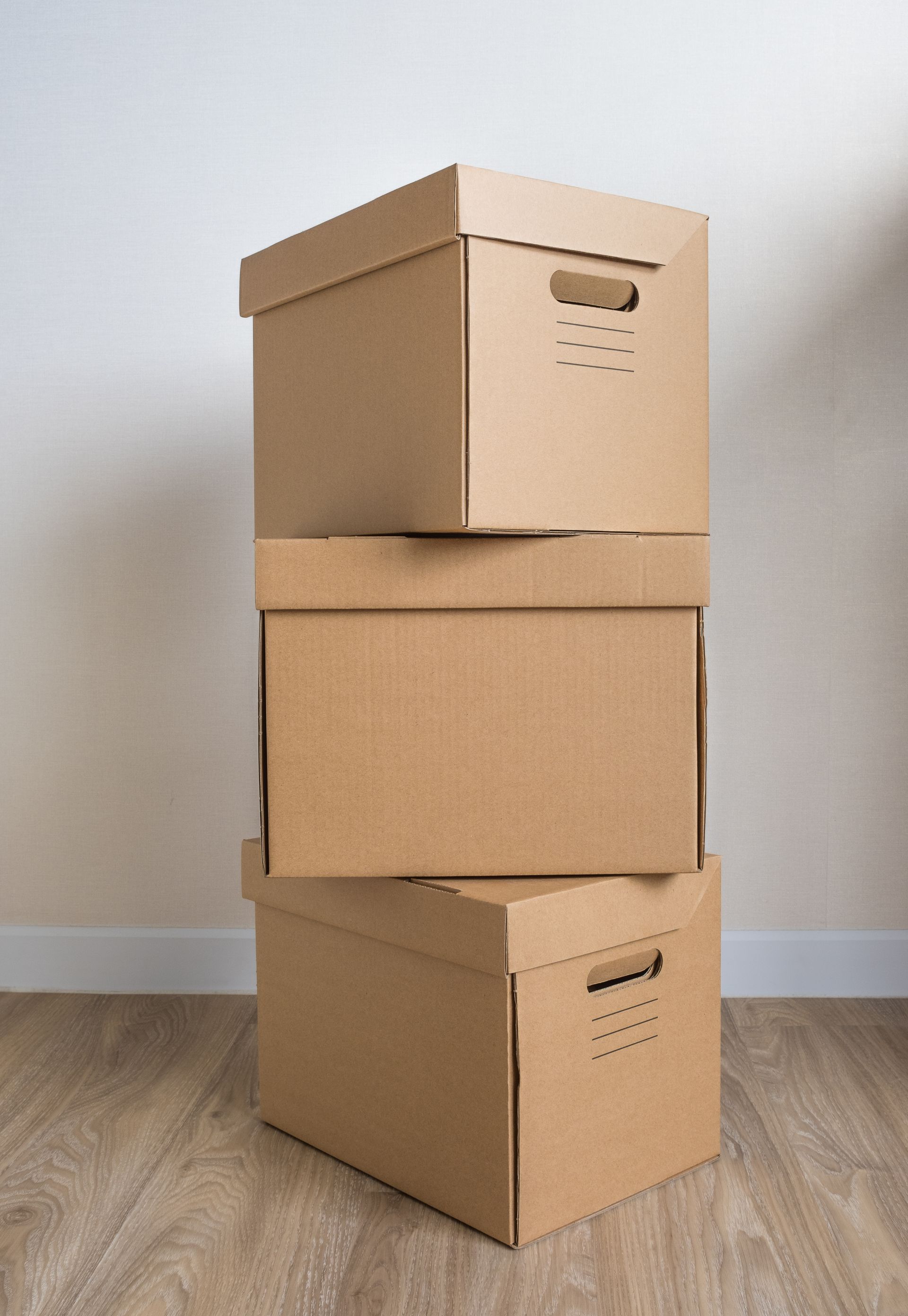 Tres cajas de cartón marrón apiladas una encima de otra, contra una pared blanca y un piso de madera.