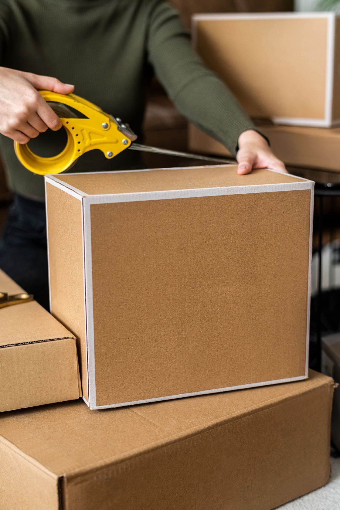 Persona sellando una caja de cartón con dispensador de cinta amarilla, cajas apiladas.