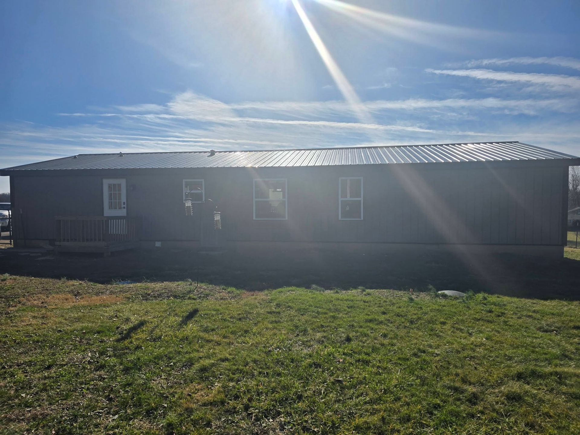 A dark grey single-story manufactured home stands in a grassy field under a bright, sunny sky with lens flare.