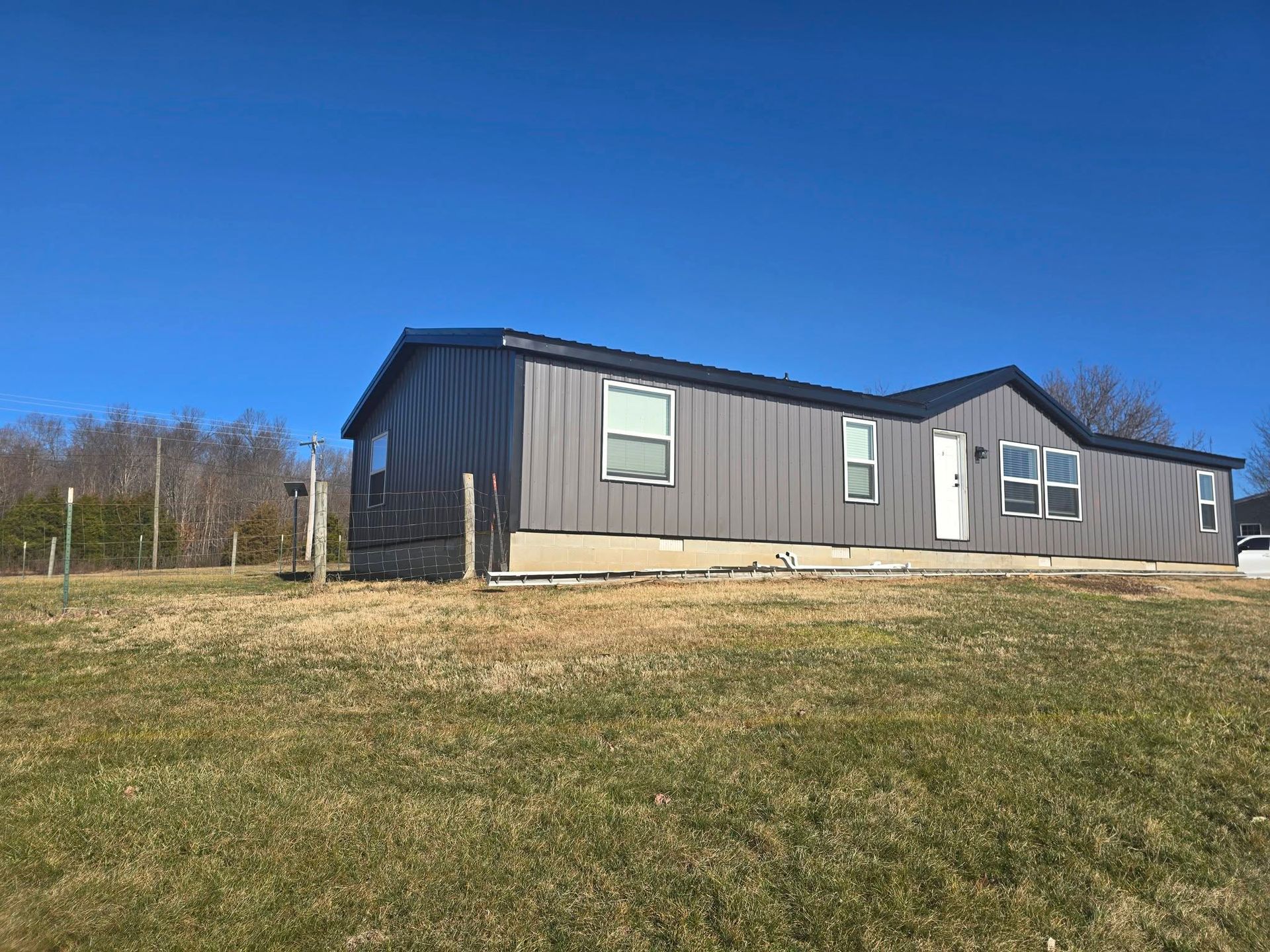 A single-story modular home with dark gray vertical siding and a black roof sits on a grassy hill under a clear blue sky.