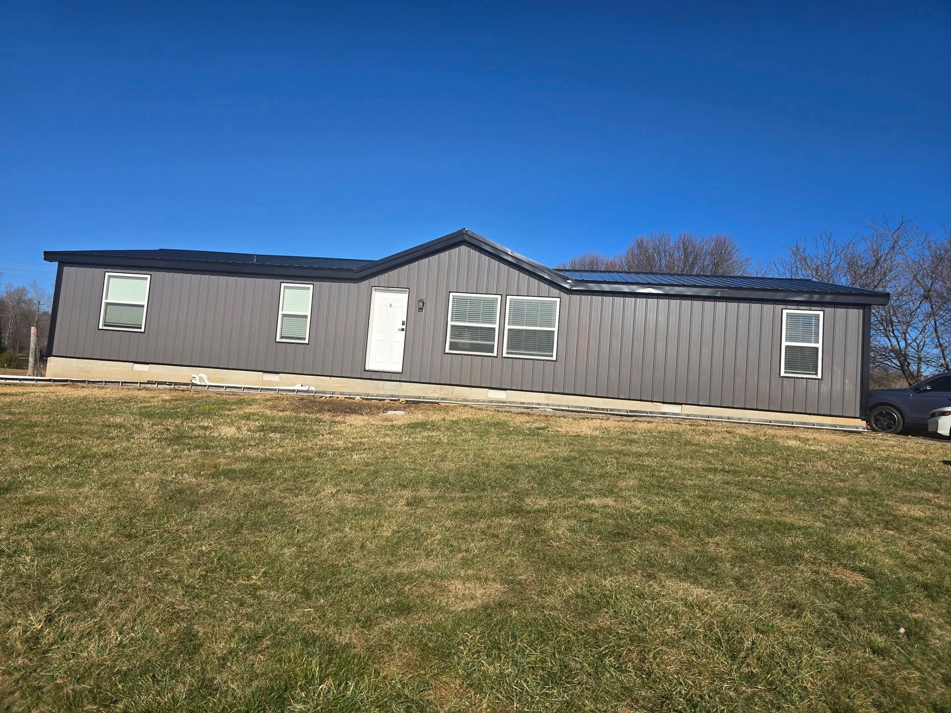 A modern gray manufactured home with a metal roof and white trim sits on a grassy lot under a clear blue sky.