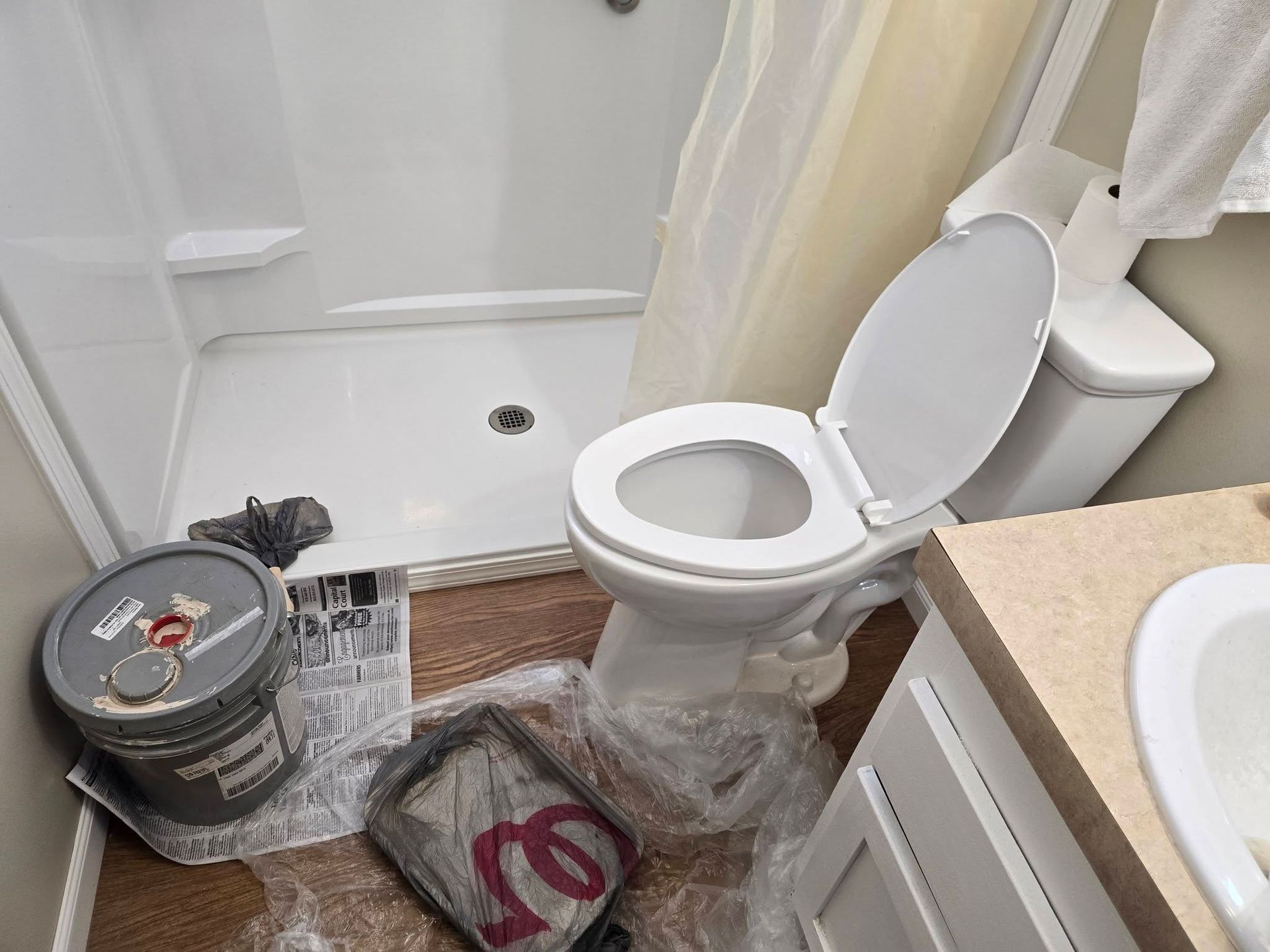 A small bathroom featuring a white toilet, a walk-in shower, and a bucket with painting supplies on the floor.