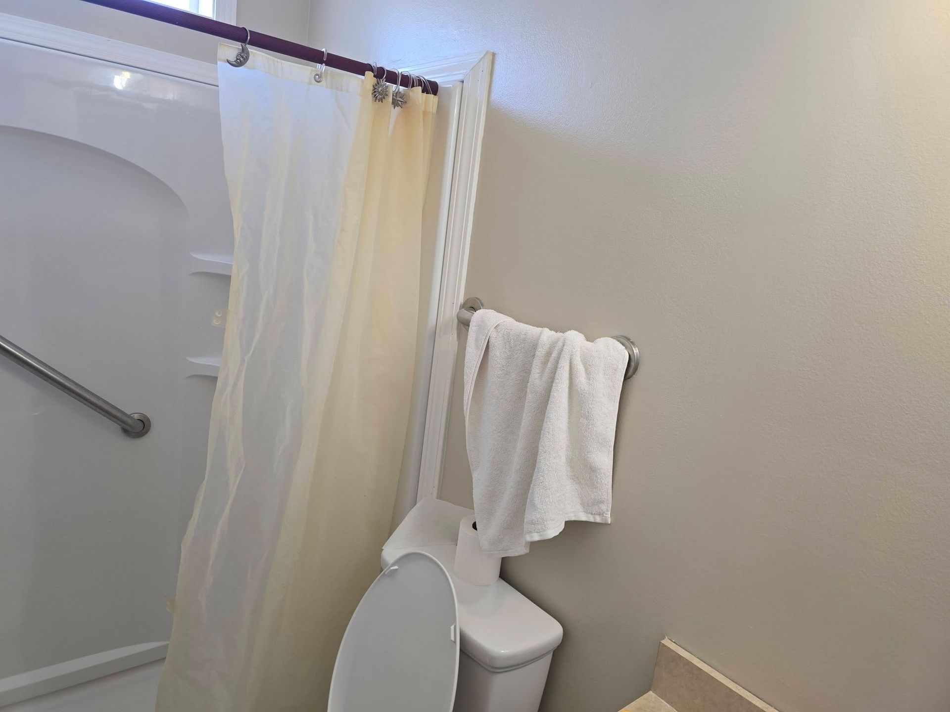 A bathroom showing a shower stall with a grab bar, a white shower curtain, and a toilet with a towel hanging on the wall.