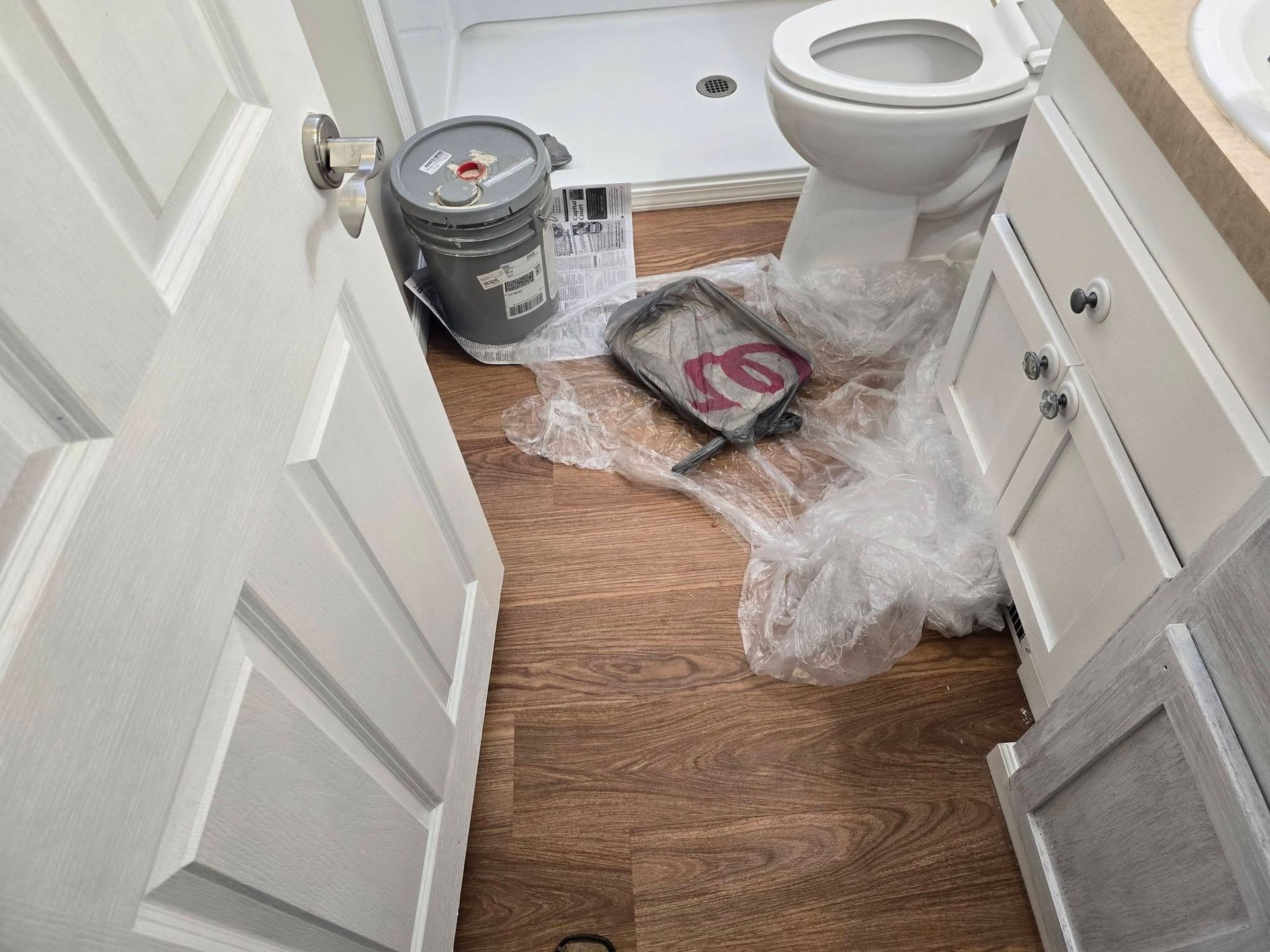 A messy bathroom under renovation featuring a paint bucket, tray, and plastic sheeting on the floor near the toilet.
