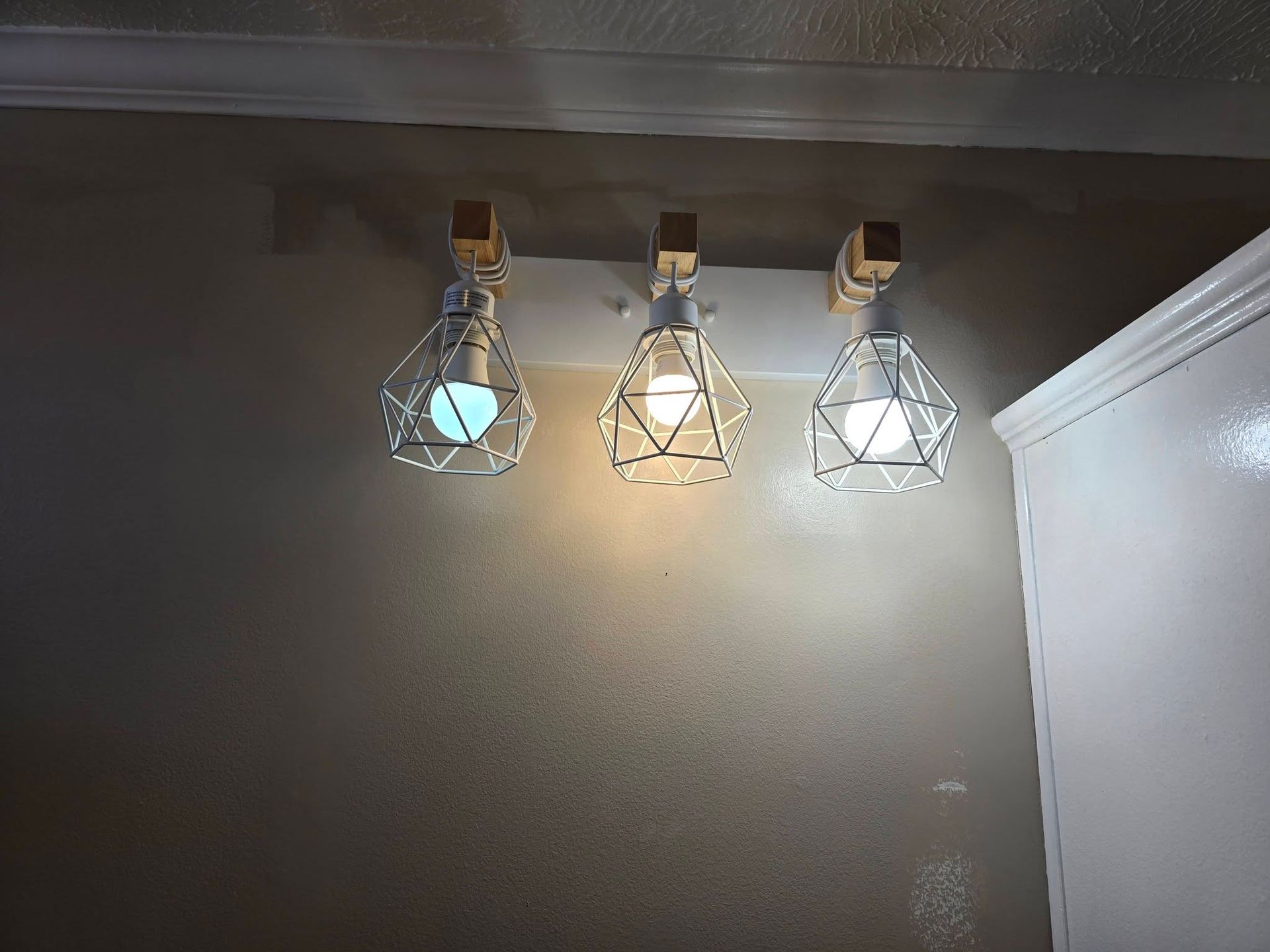 A wall-mounted light fixture with three diamond-shaped, metallic wire cages, each holding a glowing lightbulb.