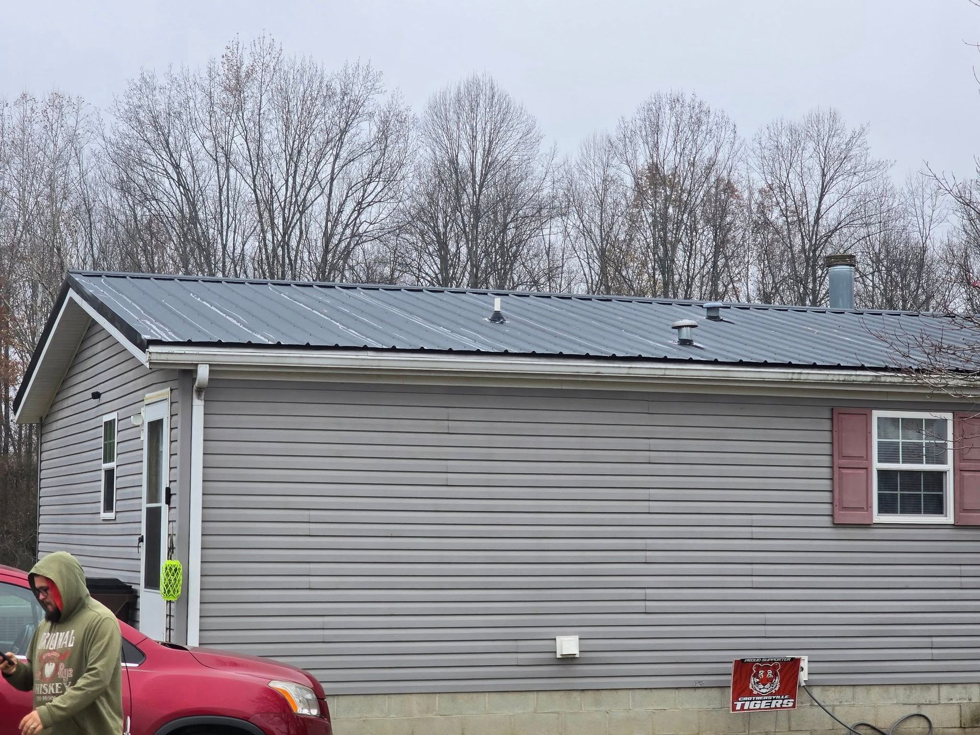 A person in a green hoodie walks past a grey vinyl-sided house with a dark metal roof and a red car parked in front.