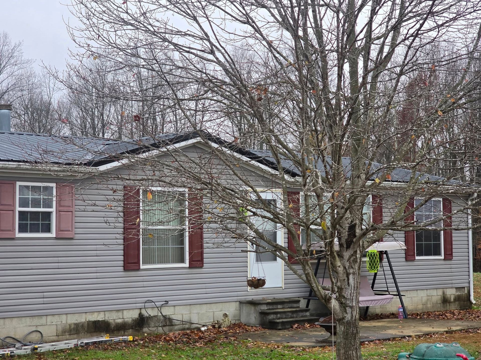 A one-story gray house with a metal roof, maroon shutters, and a bare tree in the front yard.