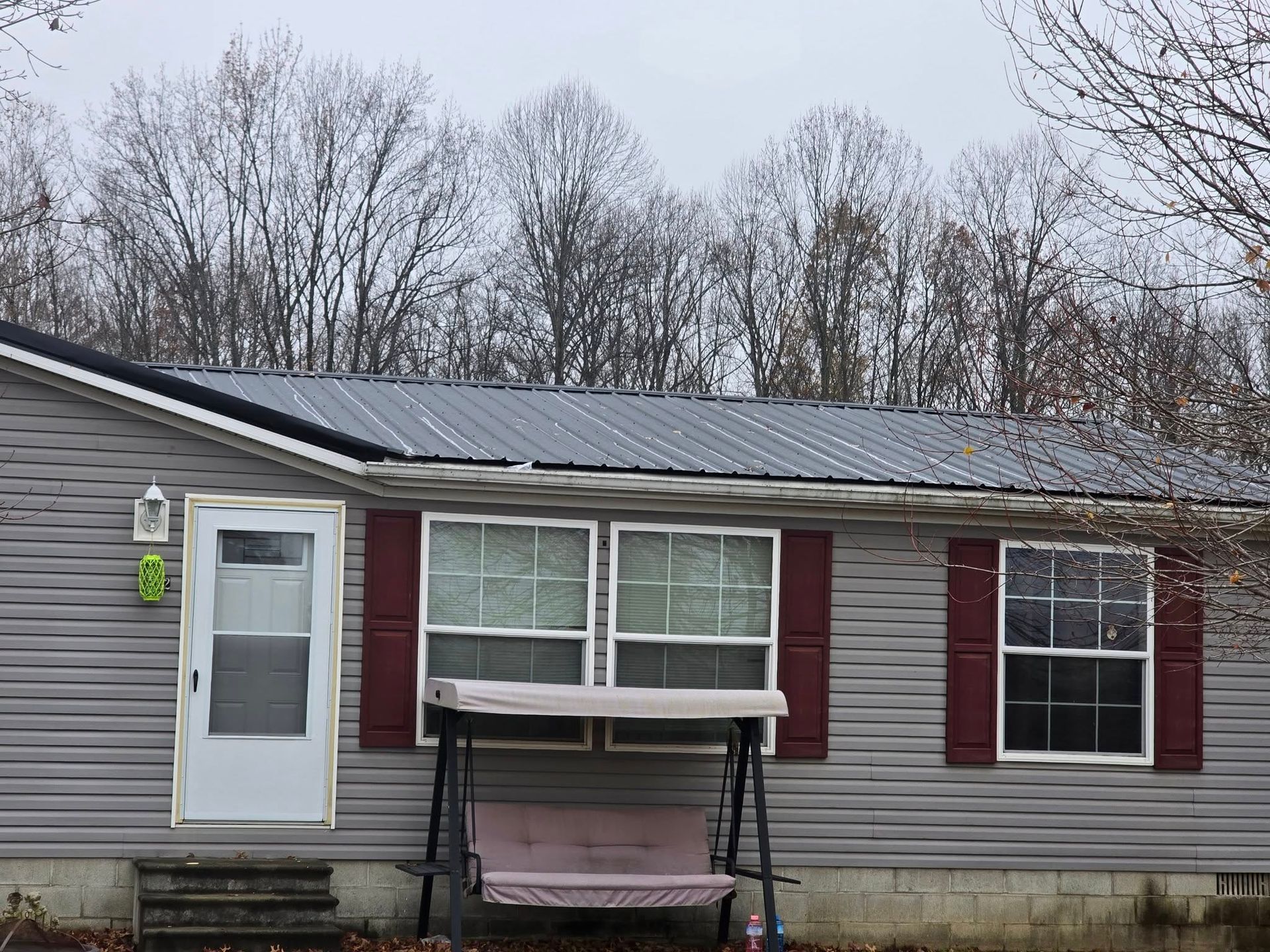 A gray, single-story house with a metal roof, white door, and maroon shutters, featuring a swing on the front porch.