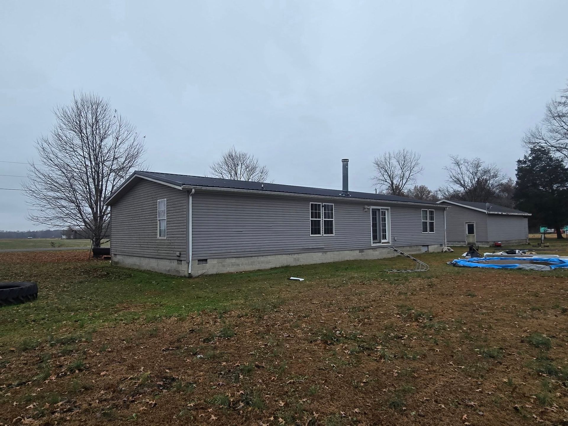 A gray manufactured home sits on a rural lot under a cloudy sky, with a blue tarp on the ground nearby.