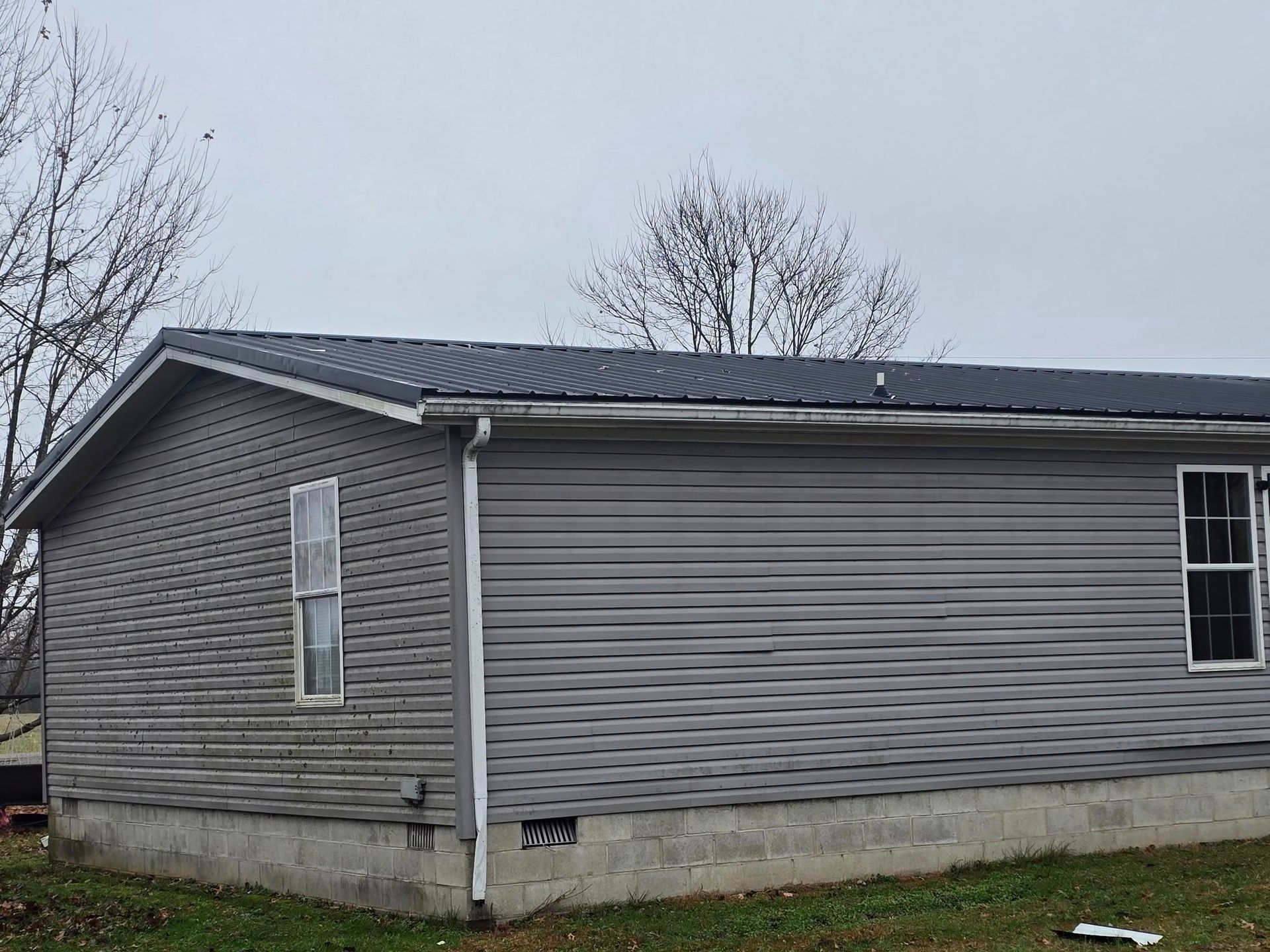 A side view of a gray vinyl-sided mobile home with a dark metal roof, set on a concrete foundation under a cloudy sky.
