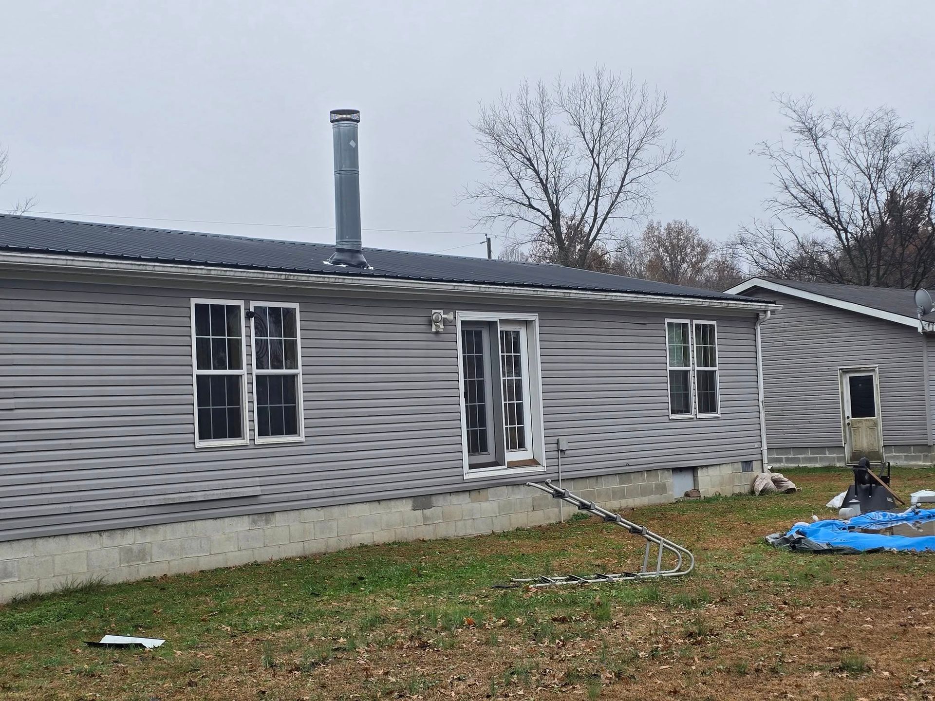 A grey, single-story manufactured home with a metal roof and chimney, situated in a yard on an overcast day.