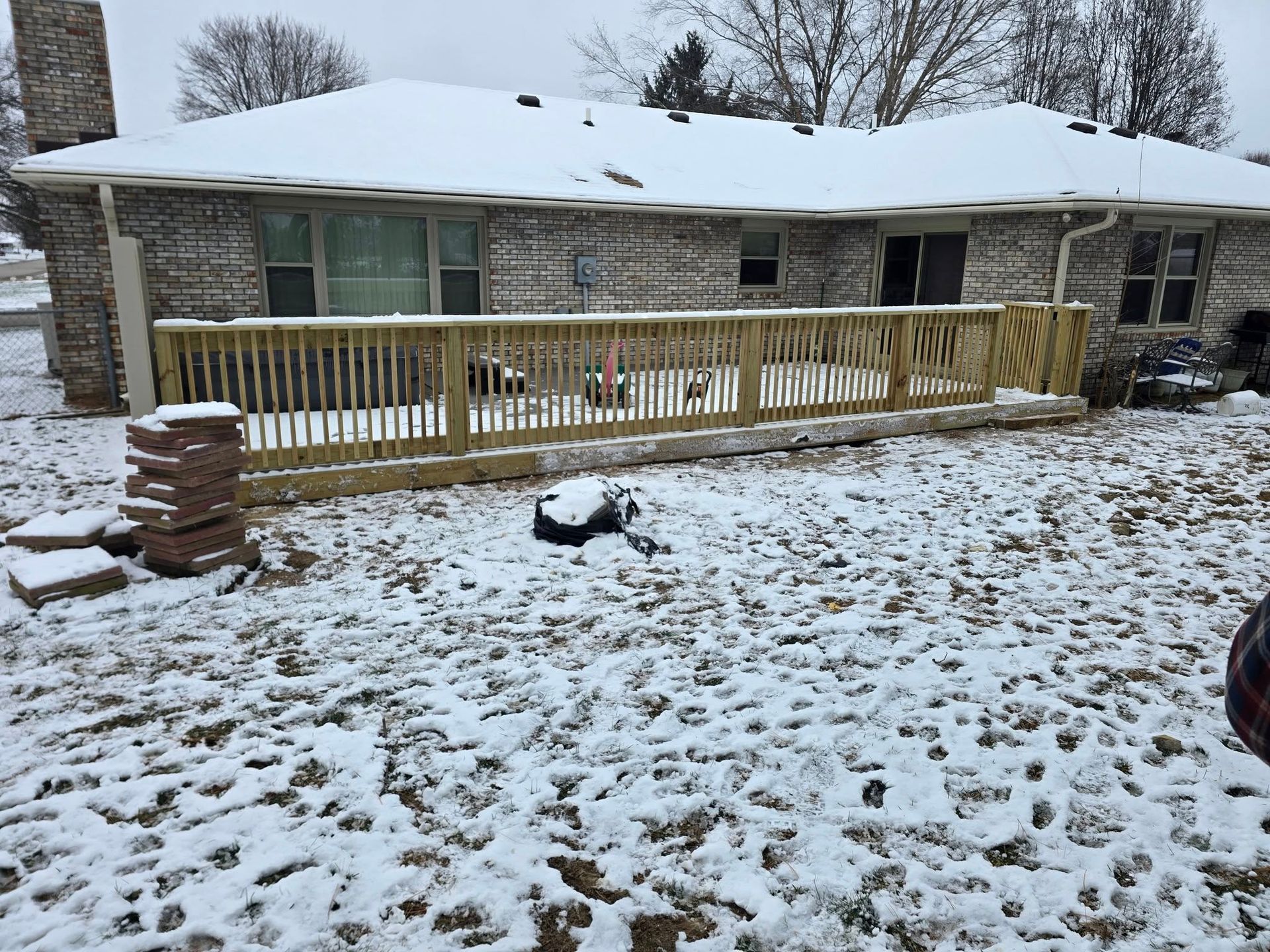 A newly built wooden deck attached to the back of a brick house, all covered in a light layer of winter snow.