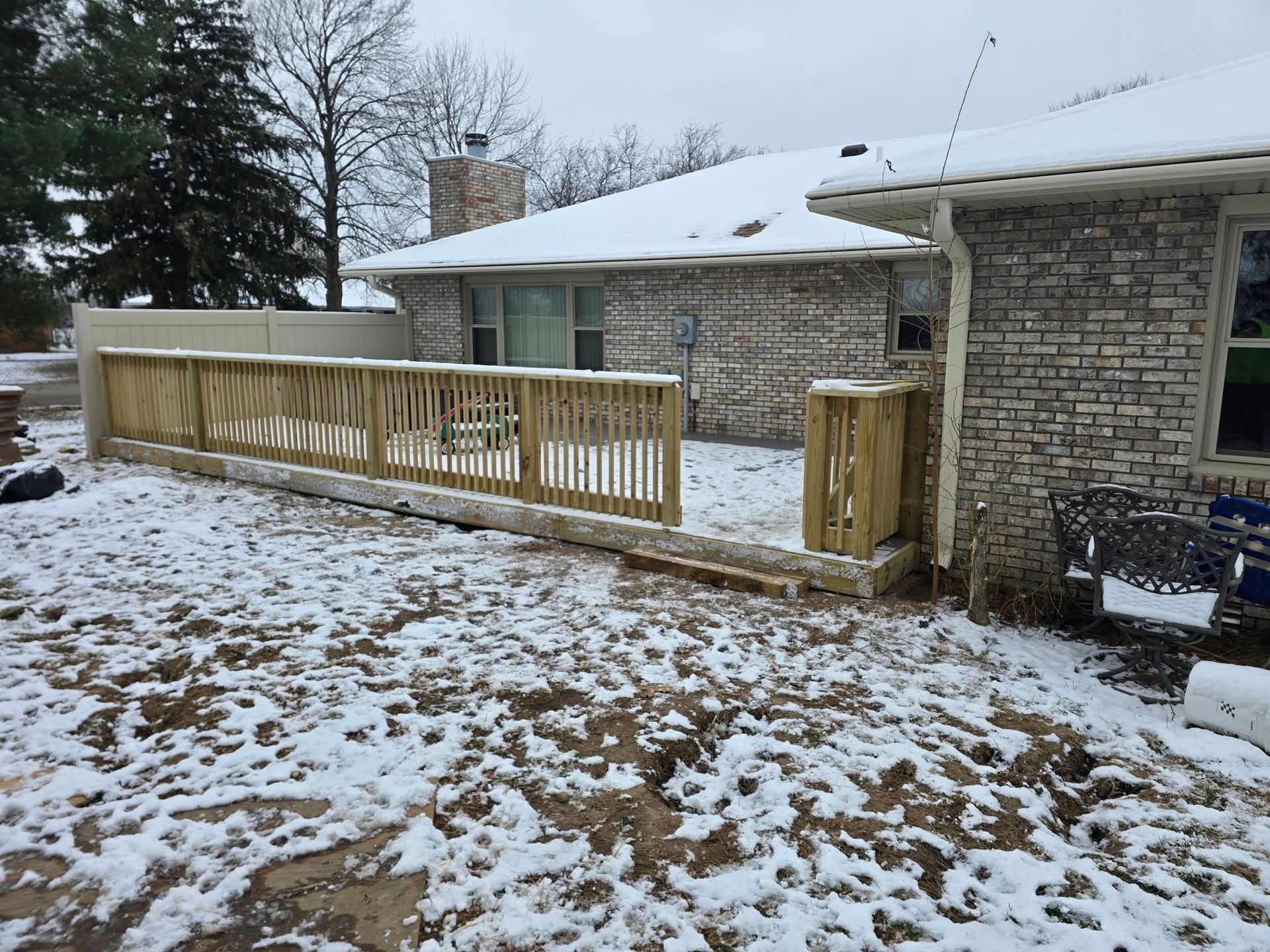 A newly built wooden deck with railings attached to the back of a brick house, both covered in a light layer of snow.