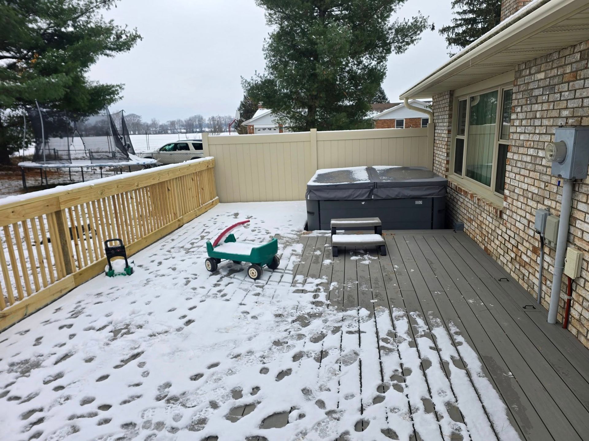 A snow-covered wooden deck featuring a hot tub, a child's toy wagon, a trampoline in the yard, and a brick house wall.