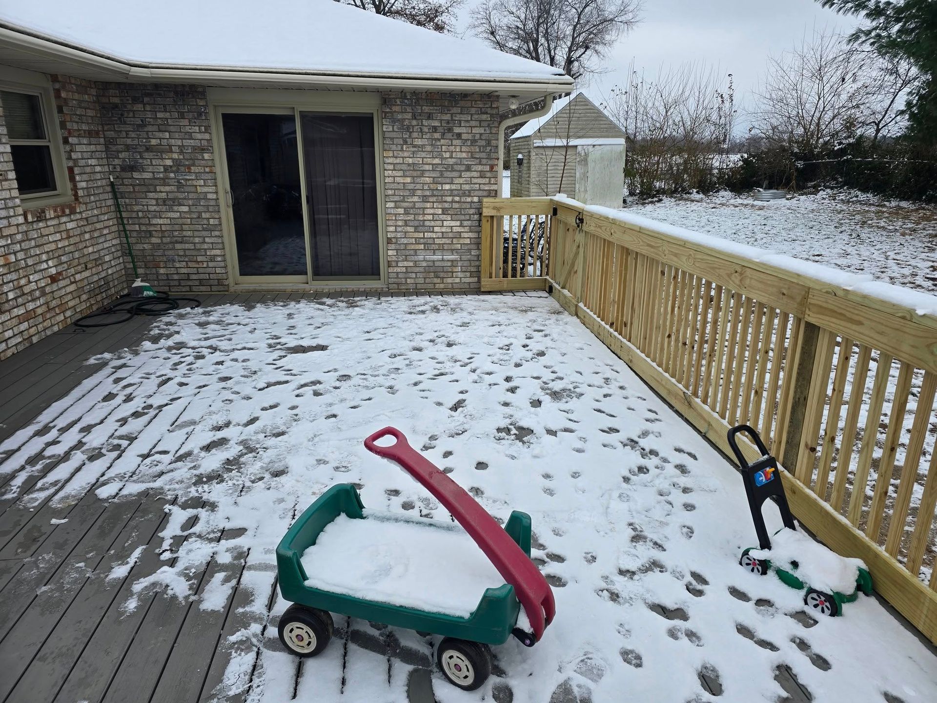 A snow-covered wooden deck outside a brick house, featuring a toy wagon and a toy lawnmower.