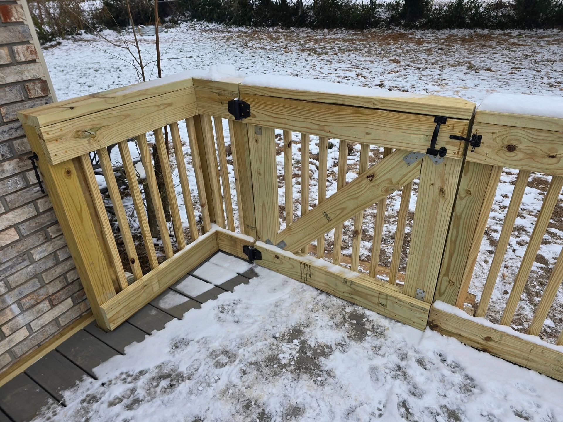 A wooden deck gate with vertical spindles, metal hinges, and a latch, installed on a deck covered in snow.
